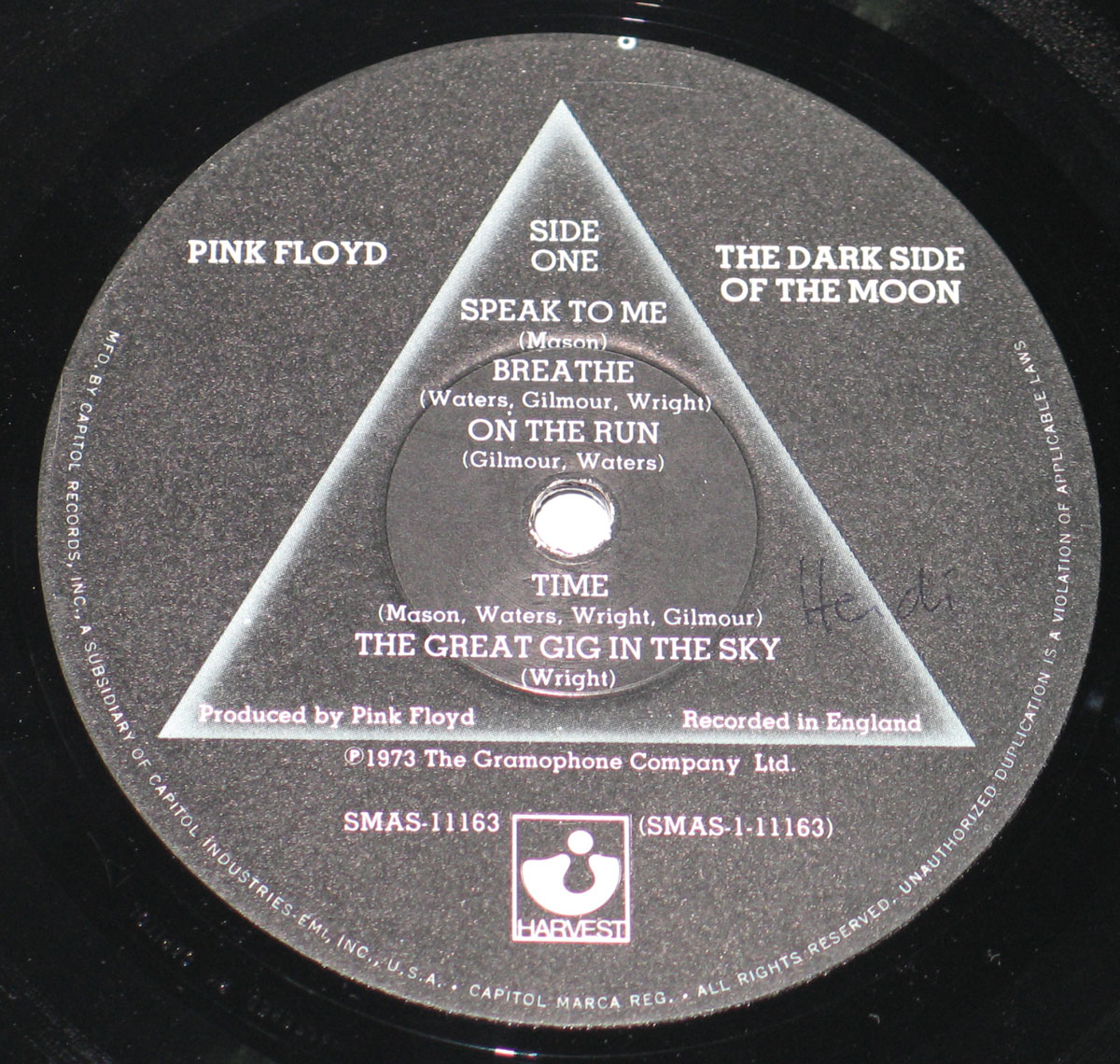 ‘Record label for Pink Floyd’s The Dark Side of the Moon, Side One. The label features a black background with a triangular prism graphic in the center, echoing the album’s iconic cover art. Song titles and composer credits are listed within the triangle. Harvest and Capitol Records logos are at the bottom, with catalog number SMAS-11163. Text around the edge details manufacturing information, stating it was made by Capitol Records in the U.S.’